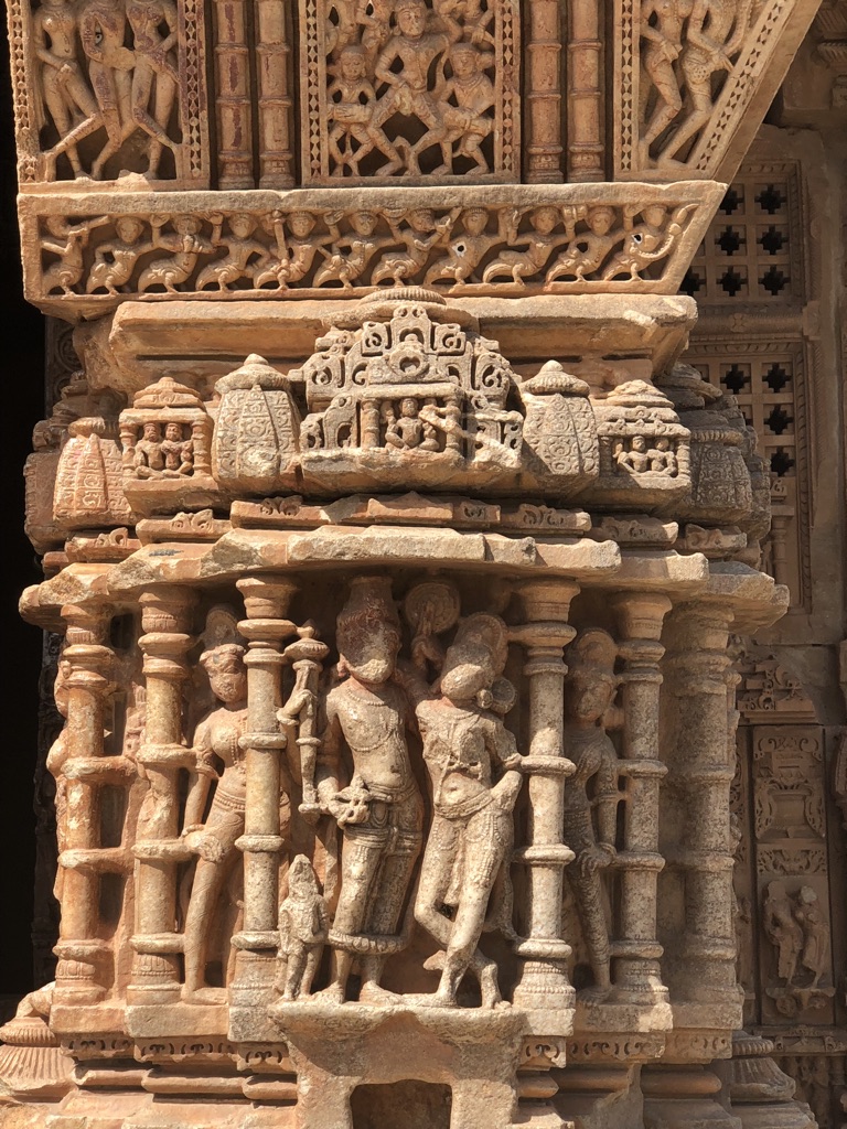 Close-up of intricate stone carvings on an ancient temple facade depicting human figures, floral patterns, and ornate architectural elements. The weathered, detailed sculptures are framed by pillars and arches, featuring mythological scenes. Bright sunlight enhances the textures.