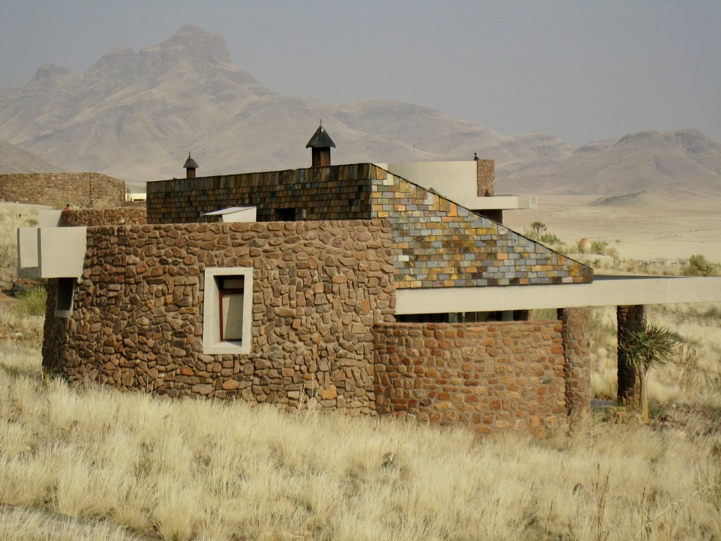 A stone-built house with a unique multicolored tiled roof, and chimneys, sits in a dry grassy field with mountains in the hazy background.