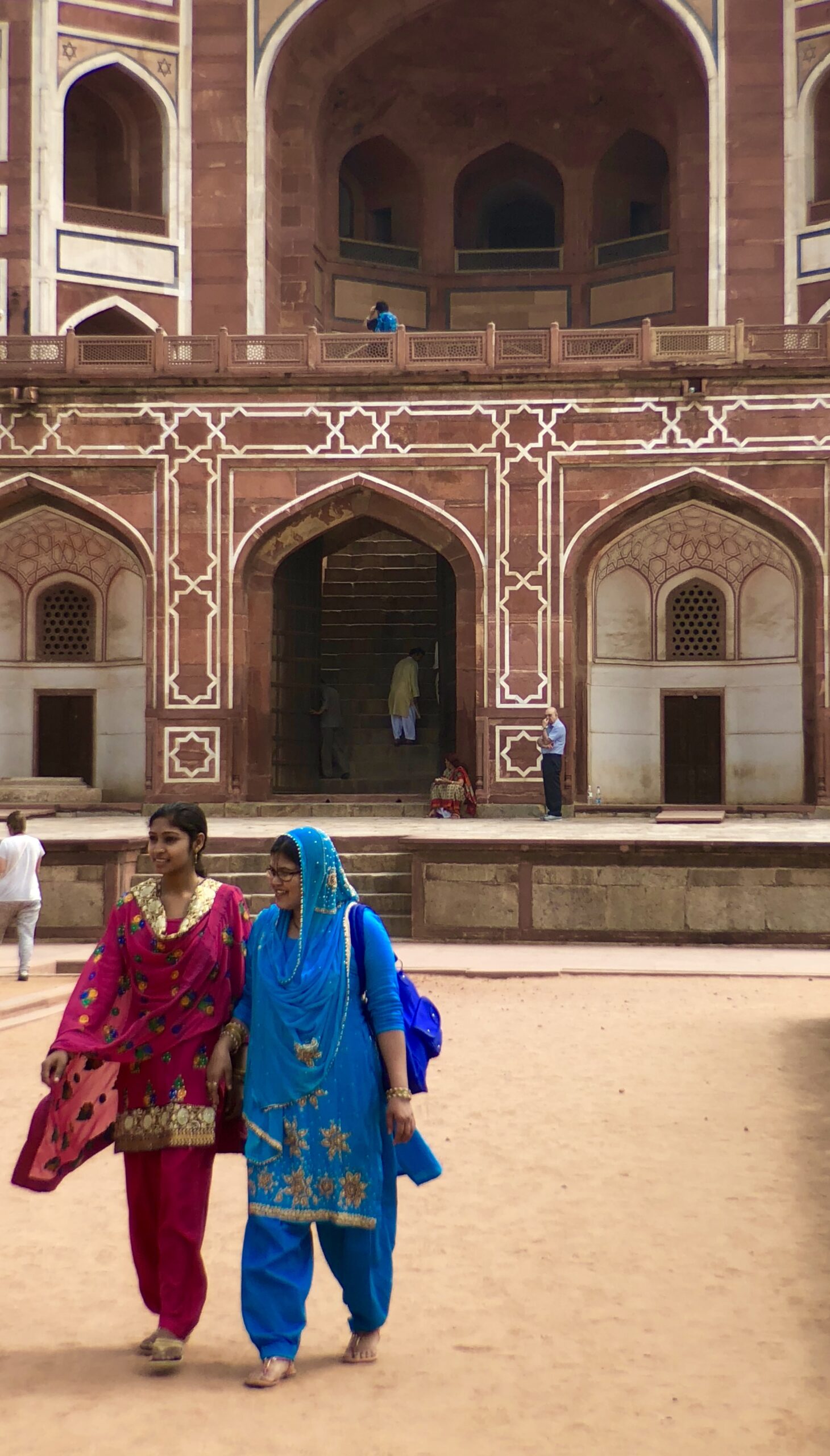 Two women wearing colorful traditional attire walk in front of a historic building with arched doorways and intricate designs. The structure is made of red and white stone. Other visitors can be seen in the background, exploring the site.