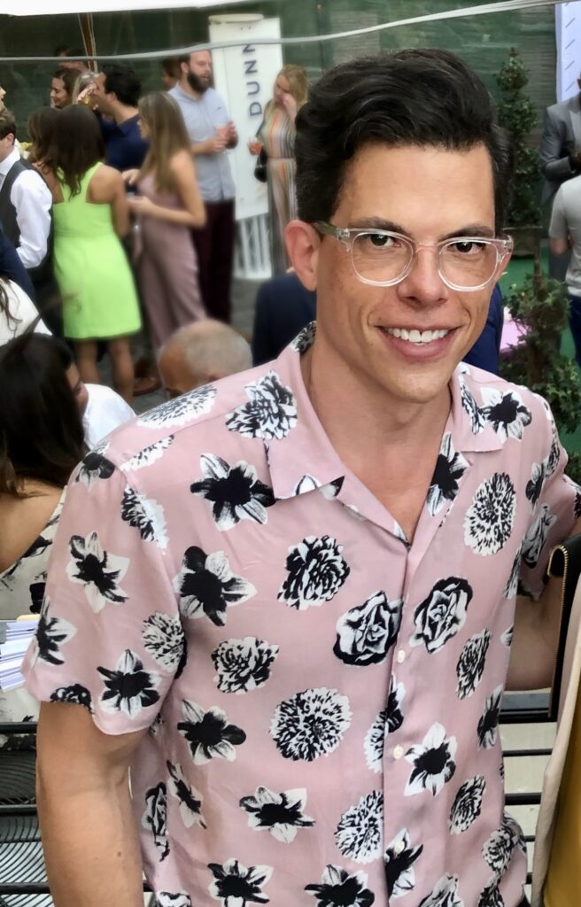 A person wearing glasses and a pink floral shirt is smiling at the camera. The background features a social gathering with people engaged in conversation. Some individuals are dressed formally, while others are in casual attire.