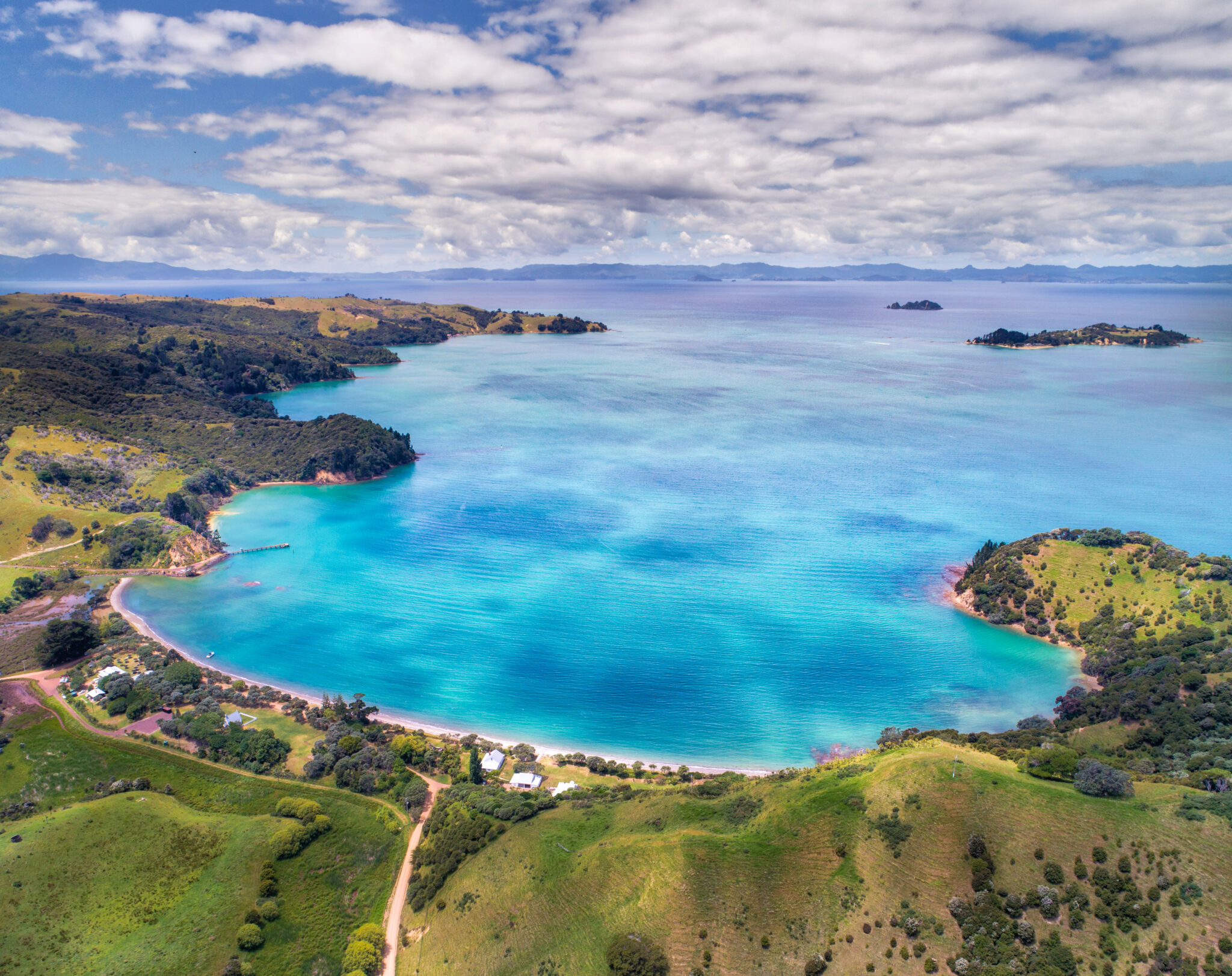 Aerial view of serene Wiheke coastal landscape featuring a turquoise bay surrounded by lush green hills and small islands in the distance. The sky is partly cloudy, adding depth to the vibrant scene. A few buildings and roads are visible near the shoreline.