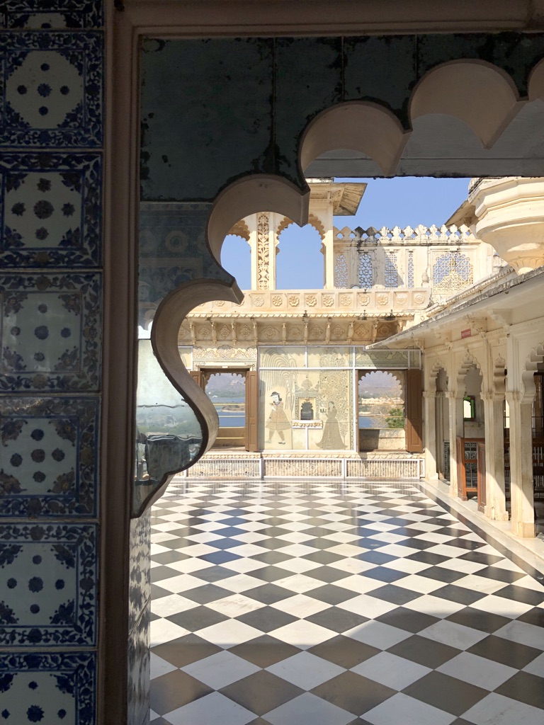 A view from an ornate arched window showcasing a courtyard with a checkered black-and-white marble floor. The surrounding walls are decorated with intricate carvings and delicate lattice work. Tiles with blue floral patterns frame the window. The sky is clear.