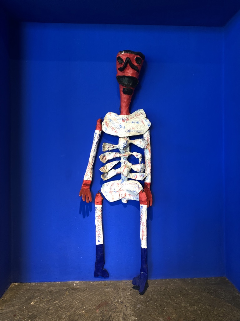 A colorful skeleton sculpture stands against a bright blue background. The skeleton has a red face and arms, with a large black mustache and blue boots. The torso and upper legs are wrapped in a white material with multicolored speckles.