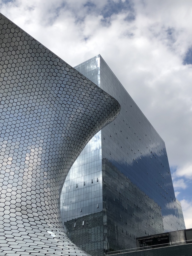 A curvaceous, hexagonal-patterned metallic building stands beside a sleek, reflective glass skyscraper under a partly cloudy sky. The two structures create a striking contrast in architectural styles, with the modern design prominently featured.