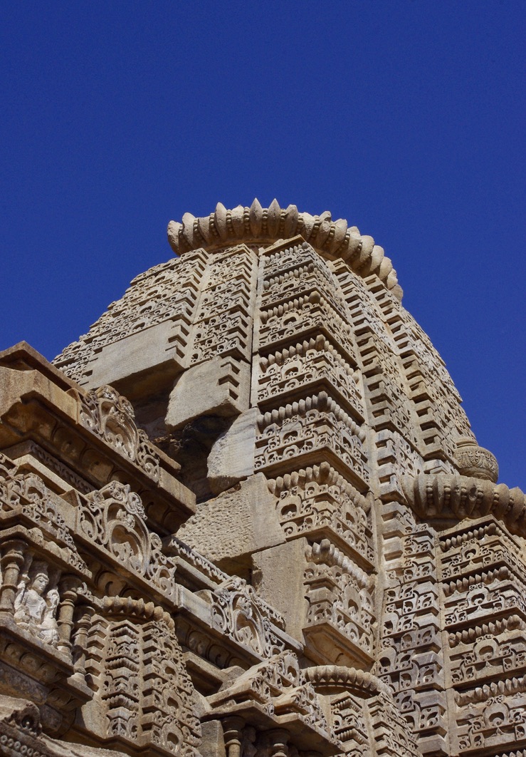 A close-up view of an intricately carved stone temple tower set against a clear blue sky. The detailed carvings feature ornate patterns and figures, showcasing artistic craftsmanship. The architectural style appears to be traditional and historical.