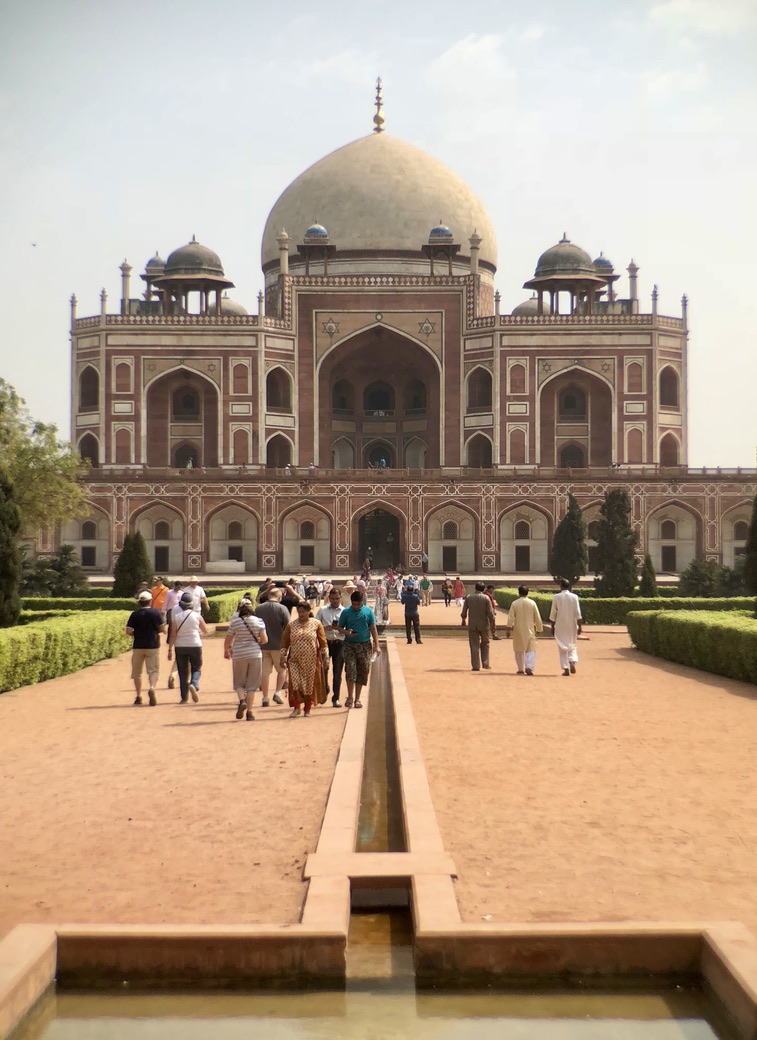A group of people walking toward Humayun's Tomb in Delhi, India. The tomb is an impressive structure with a large dome and intricate architectural details. The pathway leading to the entrance is flanked by green lawns and a narrow water channel.