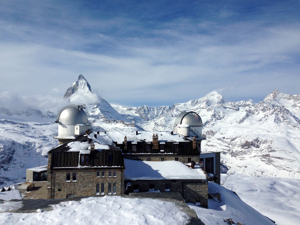 A stone observatory building with two domed telescopes sits in a snowy, mountainous landscape. The background features snow-covered peaks, including the iconic, pyramidal Matterhorn mountain under a clear blue sky.