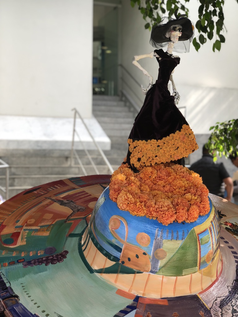 A decorative sculpture featuring a skeleton dressed in a black dress with an orange marigold skirt. The sculpture stands on a colorful base depicting abstract cityscapes and landscapes. The background shows a staircase and part of a white building.