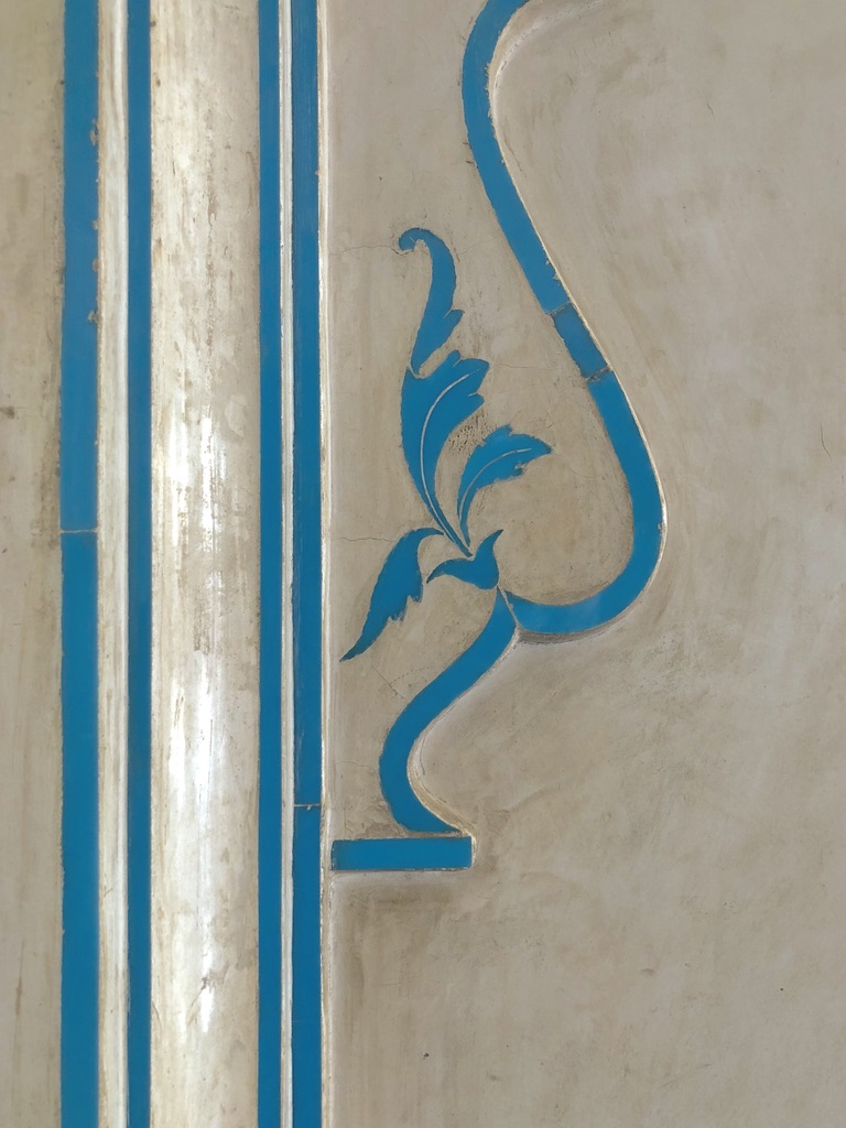 A close-up of an ornate wall design featuring blue detailing. The blue accents outline a curved, abstract shape resembling a stylized plant or leaf pattern on a light beige background. Vertical blue lines run parallel to the design on the left side.
