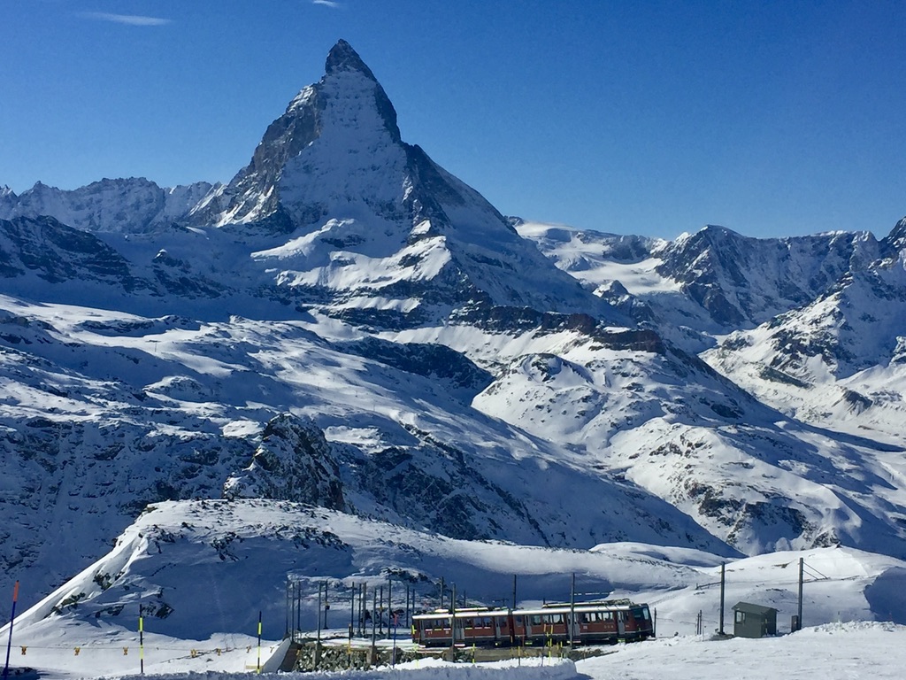 A red and white train travels through a snow-covered mountainous landscape under a clear blue sky, with the iconic Matterhorn peak prominently visible in the background. The scene is serene with vast expanses of white snow and rugged peaks.