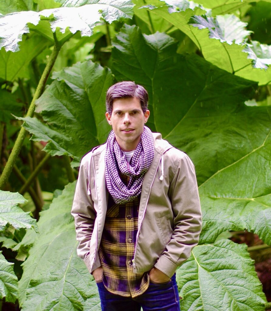 A man in a tan jacket over a checkered shirt and a scarf, stands outdoors with his hands in his pockets. He is surrounded by large green leaves, creating a lush, natural background.