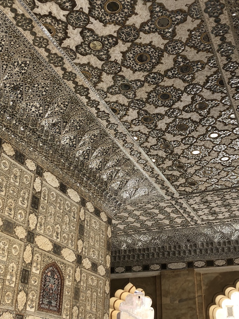 Ornate interior of the Sheesh Mahal in Amber Fort, Jaipur, showcasing intricate mirror and glass work on the walls and ceiling, with a complex mosaic pattern. The craftsmanship reflects Mughal-era architecture and design elements.