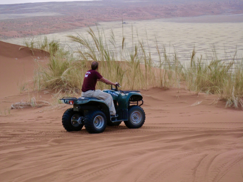 A person riding a blue quad bike on a sandy dune covered with sparse green grass, overlooking a vast, lake. the sky is overcast.