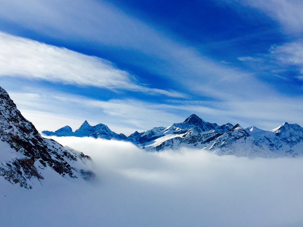 Snow-covered mountain peaks stand under a vibrant blue sky with wispy clouds. A blanket of fog or low clouds rests in the valley between the mountains, creating a striking contrast with the rugged ridges cutting through the serene atmosphere.