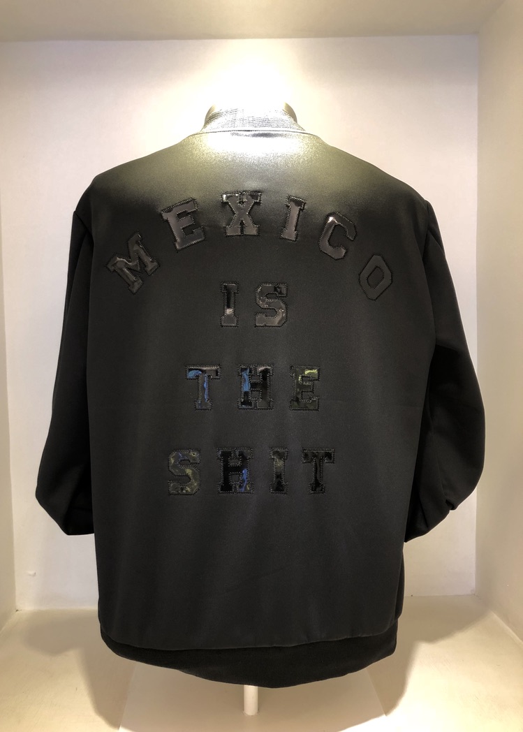 A black jacket is displayed on a white background. The back of the jacket features the text "MEXICO IS THE SHIT" in capital letters. The text is stitched in a bold, textured style. The jacket has a simple, sleek design.