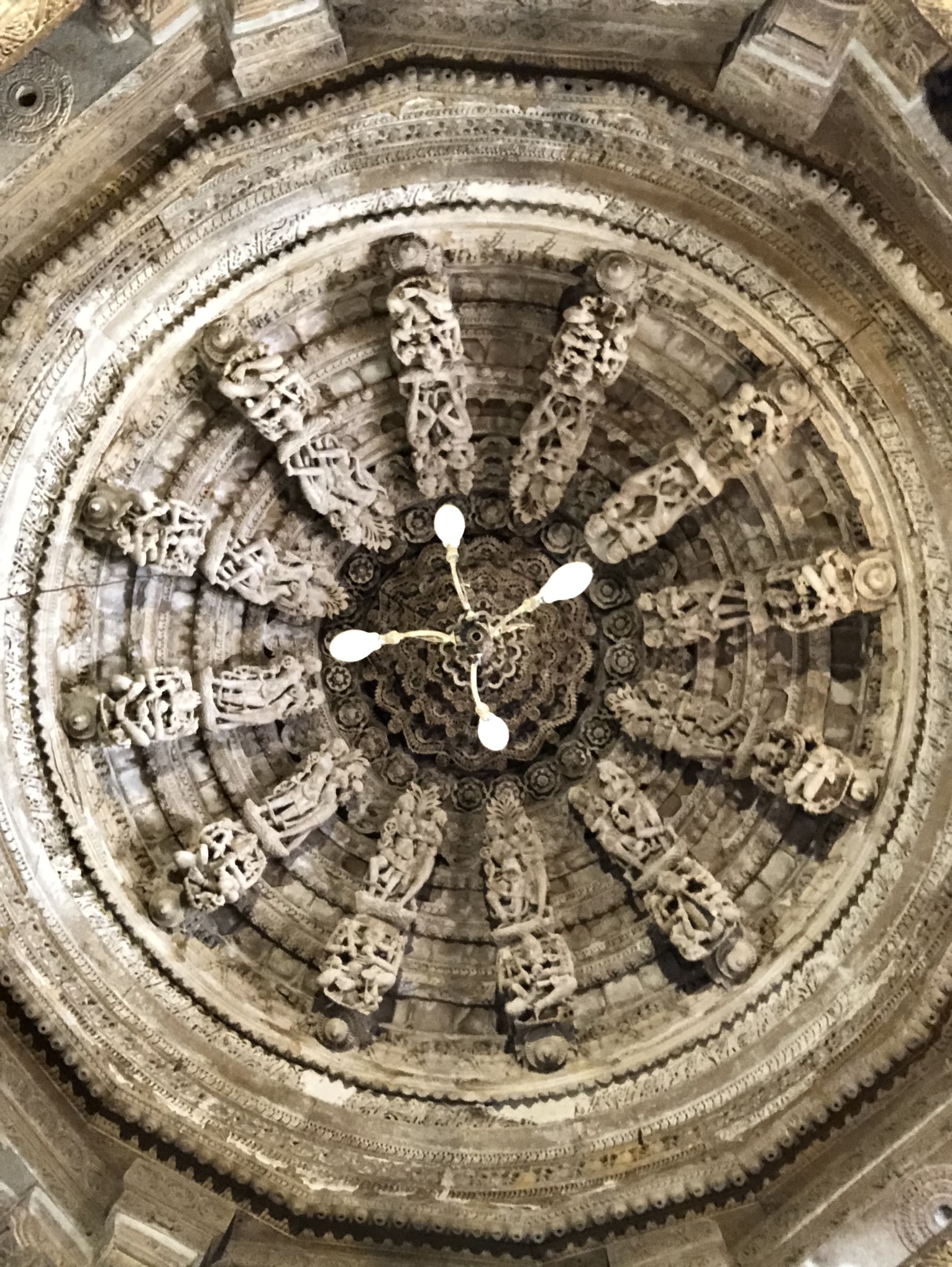 This image shows the intricately carved ceiling of an ancient temple. The circular design features detailed stone sculptures radiating outwards, and a chandelier with white lights hangs from the center. The carvings depict various figures and patterns.