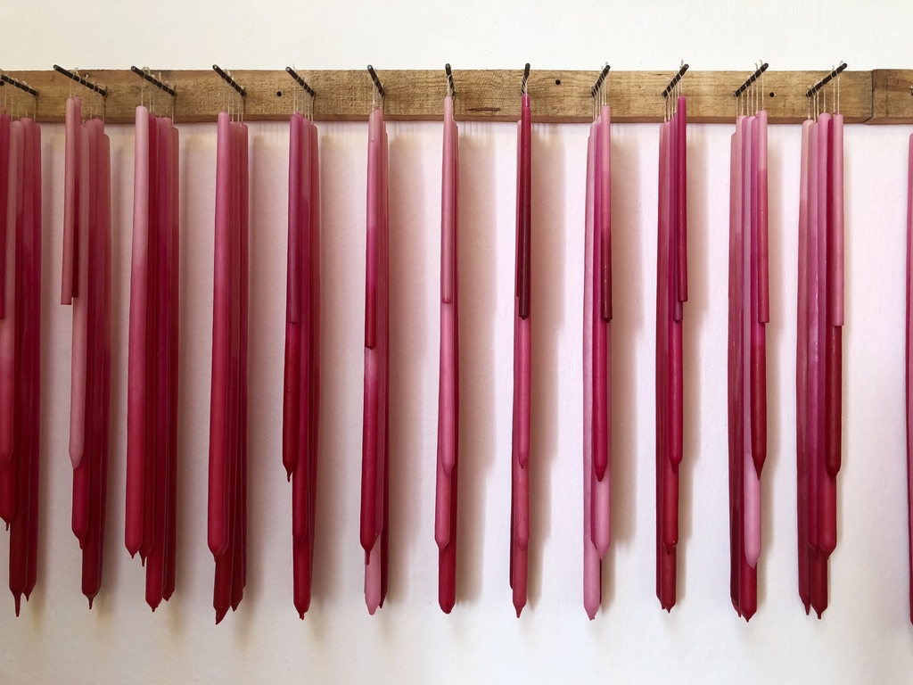 A row of pink and red candles in varying shades hangs vertically from a wooden rack mounted on a white wall. The candles are evenly spaced and elongated with a rustic appearance.