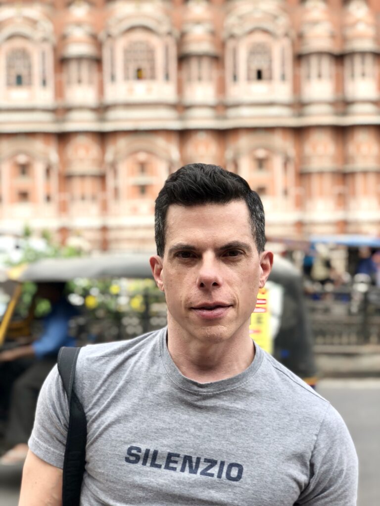 A man with short dark hair stands in front of a historic building with intricate architectural details. He is wearing a gray t-shirt that says "SILENZIO" and carrying a black shoulder bag. The background includes an auto-rickshaw and blurred pedestrians.