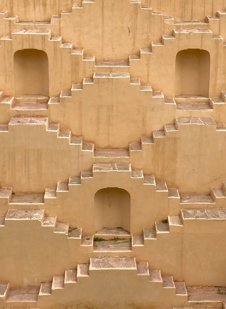 Symmetrical, beige stone staircase structure with multiple landings, creating a geometric, stepwell-like pattern. Each section has a recessed arched doorway at the top and alternating connected steps going up and down. The stone has a weathered appearance.