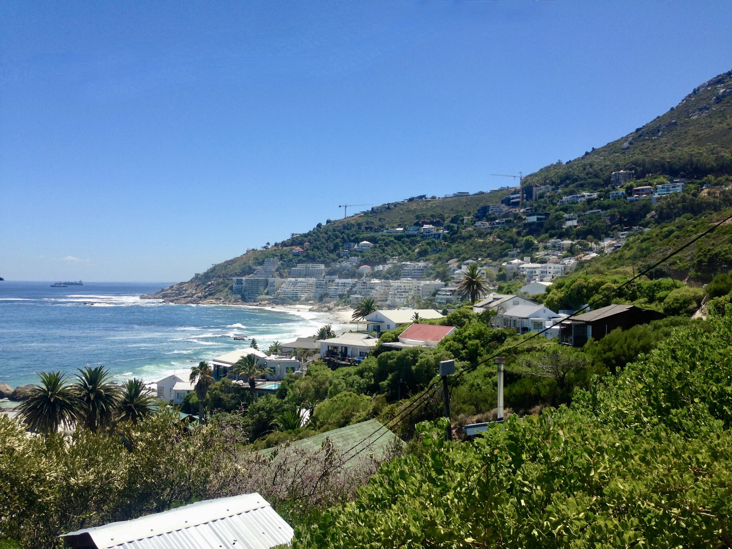 Scenic coastal view of a town nestled between lush green hills and the ocean. The hillside is dotted with houses and vegetation, with a beach stretching along the shore. Clear blue sky and calm sea visible in the background.