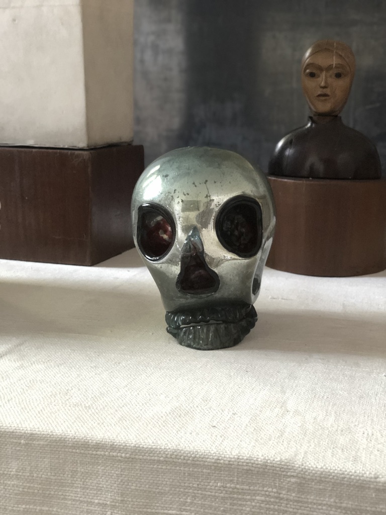 A small metallic skull sculpture with darkened eye sockets sits on a white surface. In the background, there is a wooden block with a small statue featuring a humanoid figure with a serene expression.