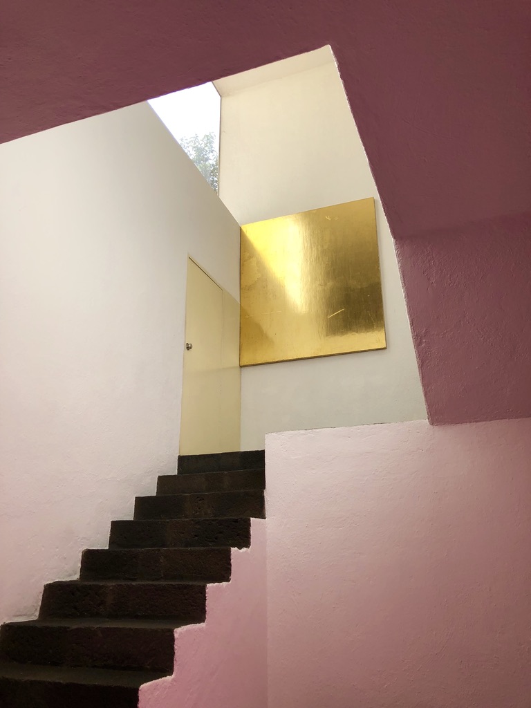 A staircase with dark steps and pink walls leads up to a higher level. A large golden square artwork hangs on the white wall at the top of the stairs. Light streams in from a skylight above, illuminating the space.