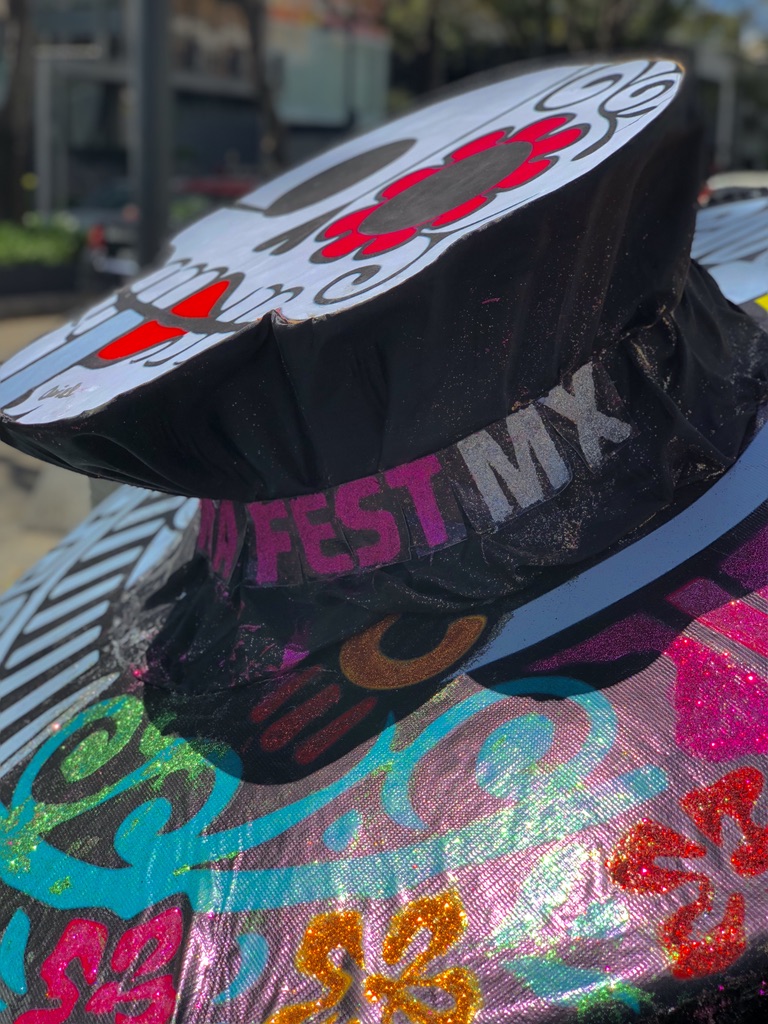 Close-up of a colorful, decorative object with intricate patterns and floral designs. The object has "FEST MX" written on it, indicating a festival or celebration. Background shows a hint of greenery and possibly an outdoor setup in a vibrant environment.