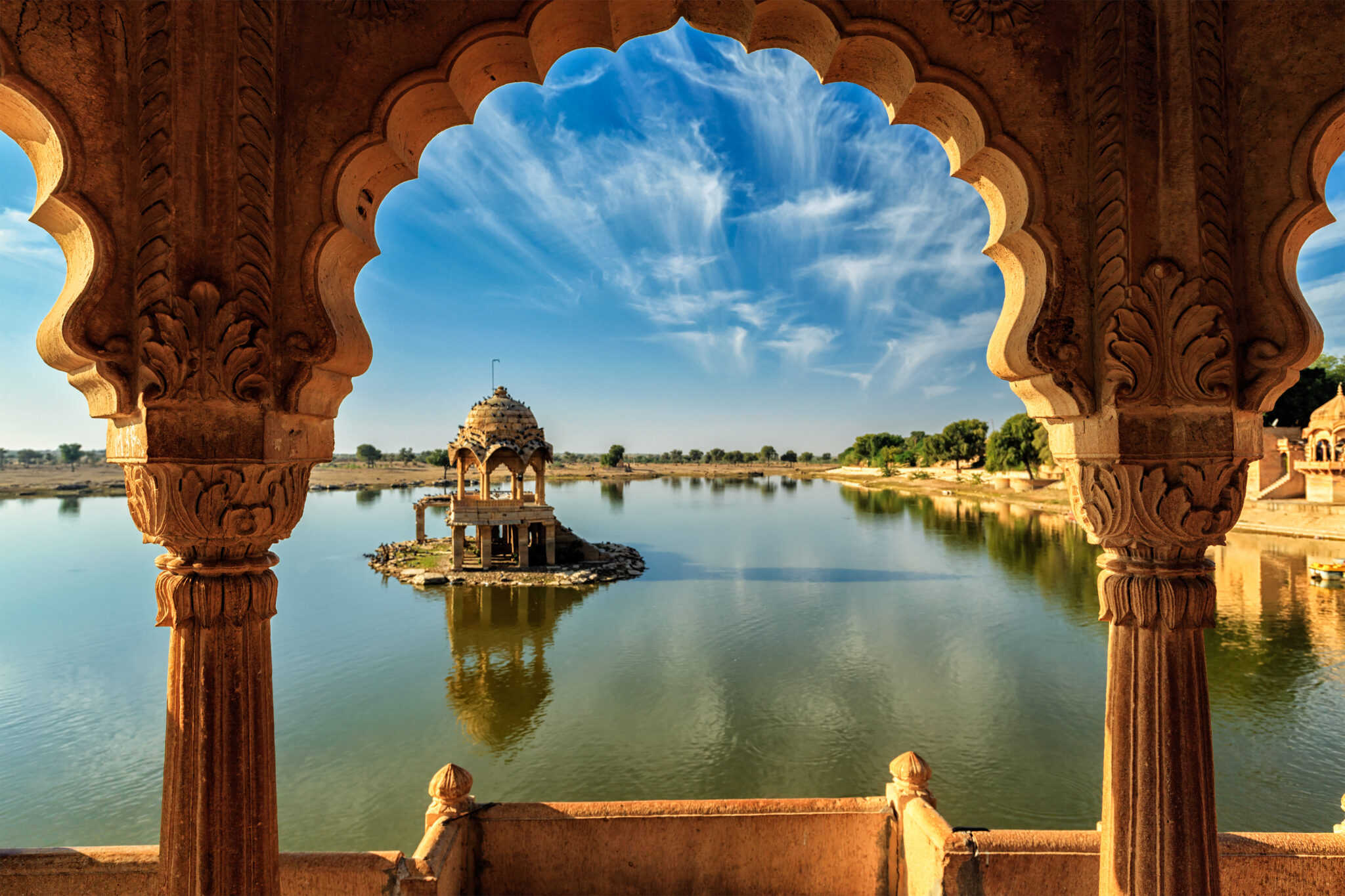 A scenic view of a historic pavilion with intricate carvings, standing on a small island, surrounded by a serene lake reflecting the blue sky and clouds. The photo is framed by the arch of a larger structure, enhancing the overall architectural beauty.