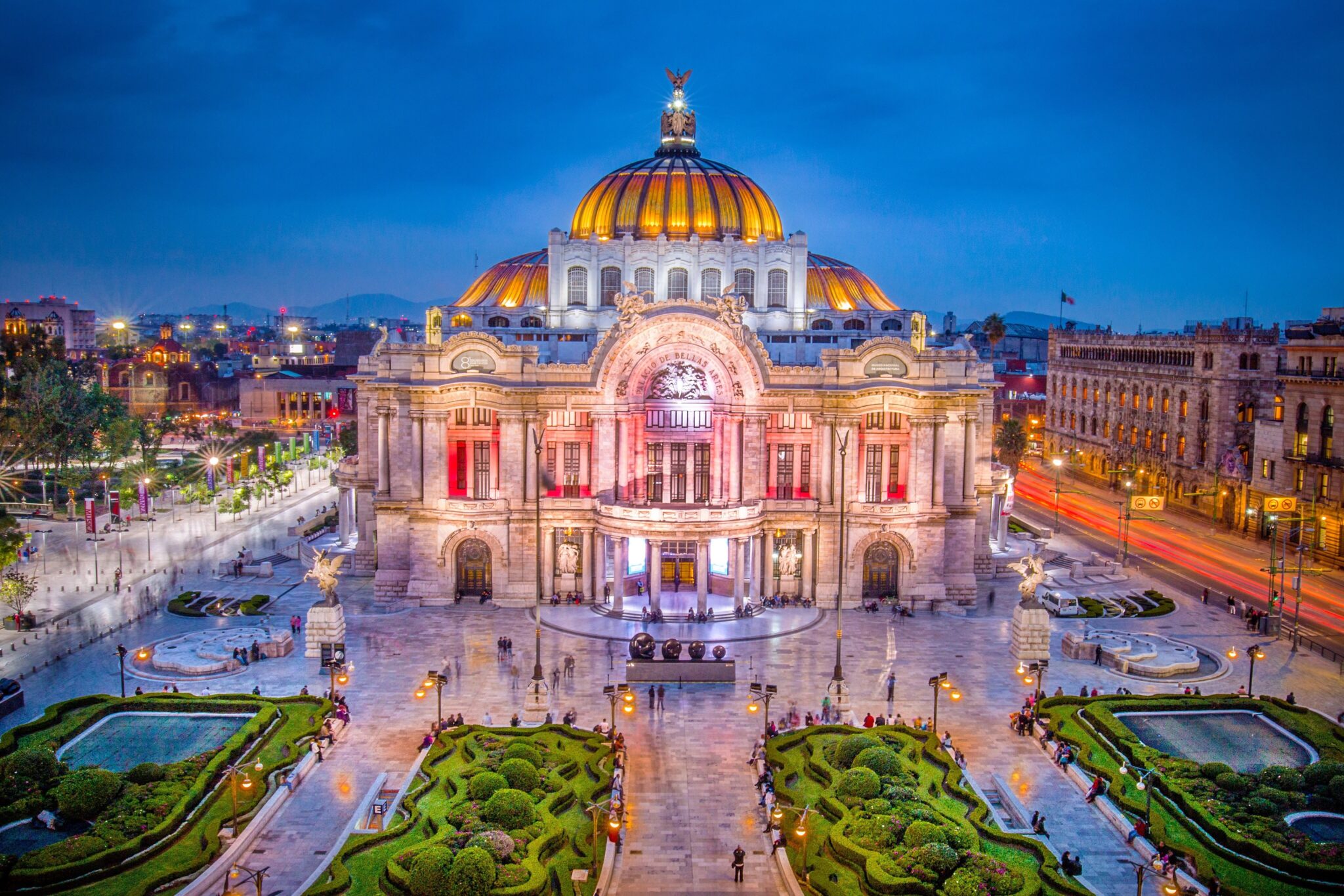 Aerial view of the Palacio de Bellas Artes (Palace of Fine Arts) in Mexico City, illuminated at dusk. The grand building features a distinctive yellow and orange dome, and the surrounding area includes neatly manicured gardens and bustling streets.