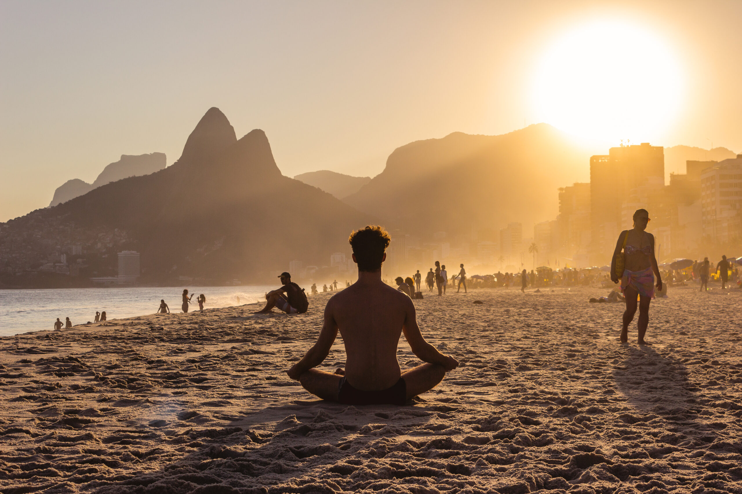 A Man sits cross-legged on a sandy Rio beach, meditating as the sun sets behind distant mountains. The silhouettes of other people walking and relaxing are visible, with the sky glowing in warm hues of orange and yellow.