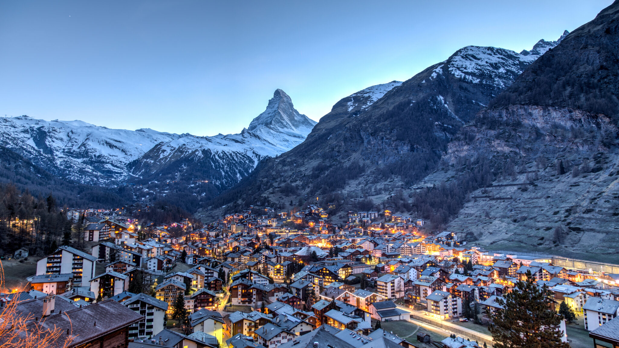 A picturesque mountain village at dusk, illuminated warmly against the backdrop of snow-capped peaks under a clear sky. The iconic Matterhorn mountain stands majestically in the background, towering over the clustered buildings below.