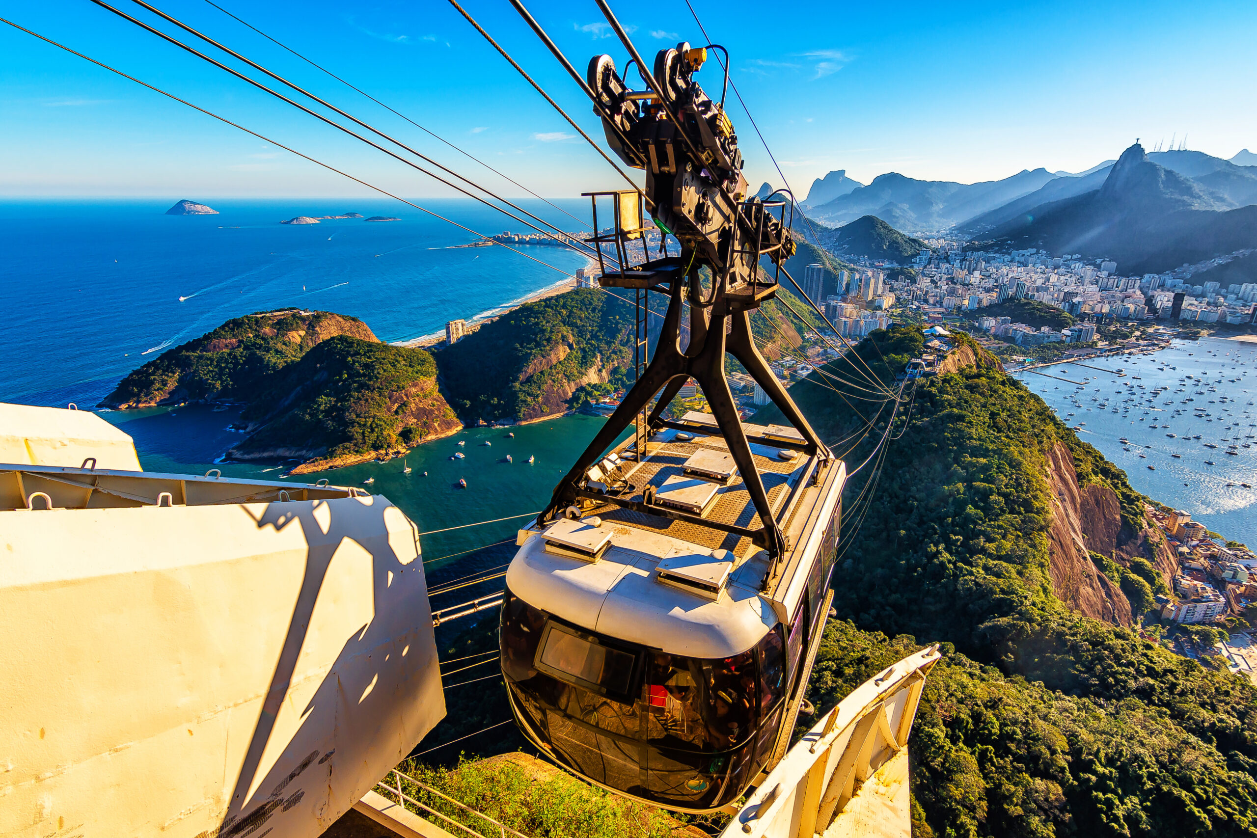 A cable car ascends toward the summit of a mountain overlooking a coastal city with sprawling urban areas, verdant hills, and expansive blue seas. Sunlight bathes the scene, highlighting the natural beauty and vibrant colors of the landscape.