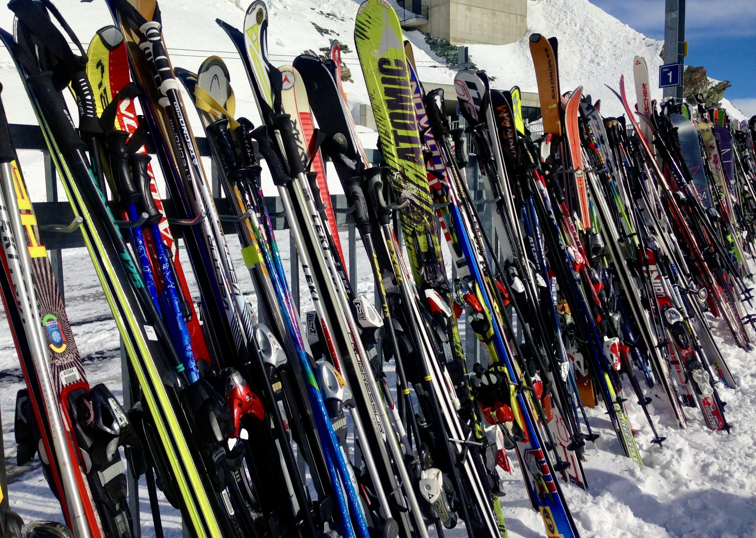 A long row of colorful skis and ski poles stacked upright in a snowy area, likely a ski resort. Snow-covered slopes and parts of a building are visible in the background under a clear blue sky.