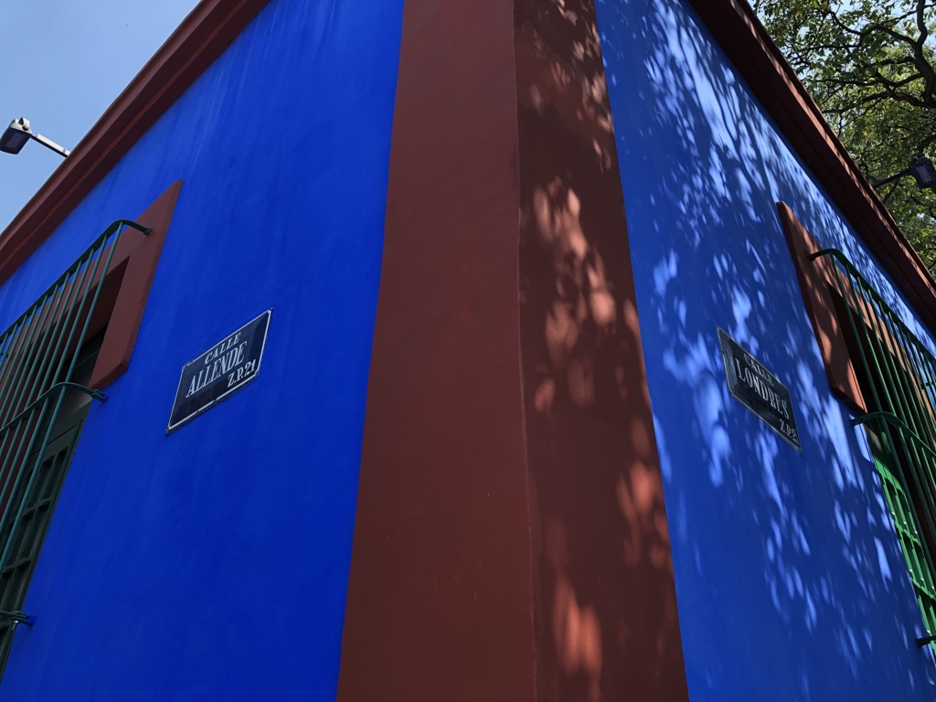 Corner of a vibrant blue and red building with green window bars. Sign on left wall reads "Calle de Allende" and "Calle de Londres" on right. Shadows of tree branches are cast on the right side. A streetlight is visible at the top left corner.