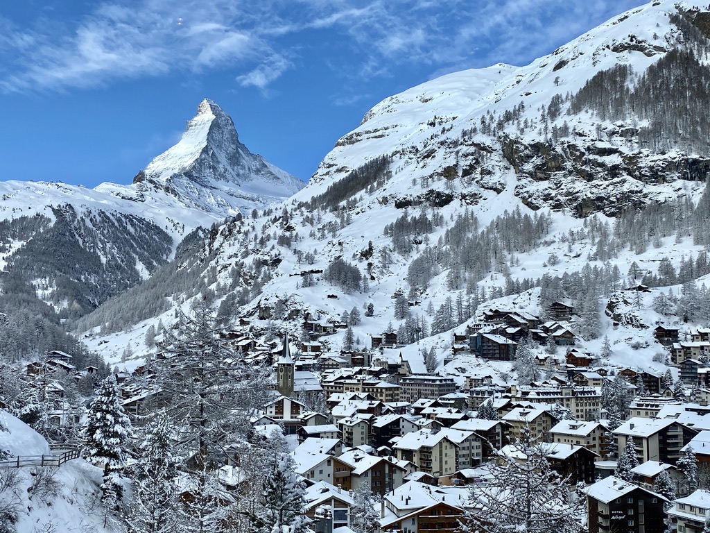 A picturesque winter scene of Zermatt, Switzerland. Snow-covered chalet-style buildings are nestled in a valley, surrounded by snow-laden trees and mountains. The iconic Matterhorn peak towers in the background under a clear blue sky.