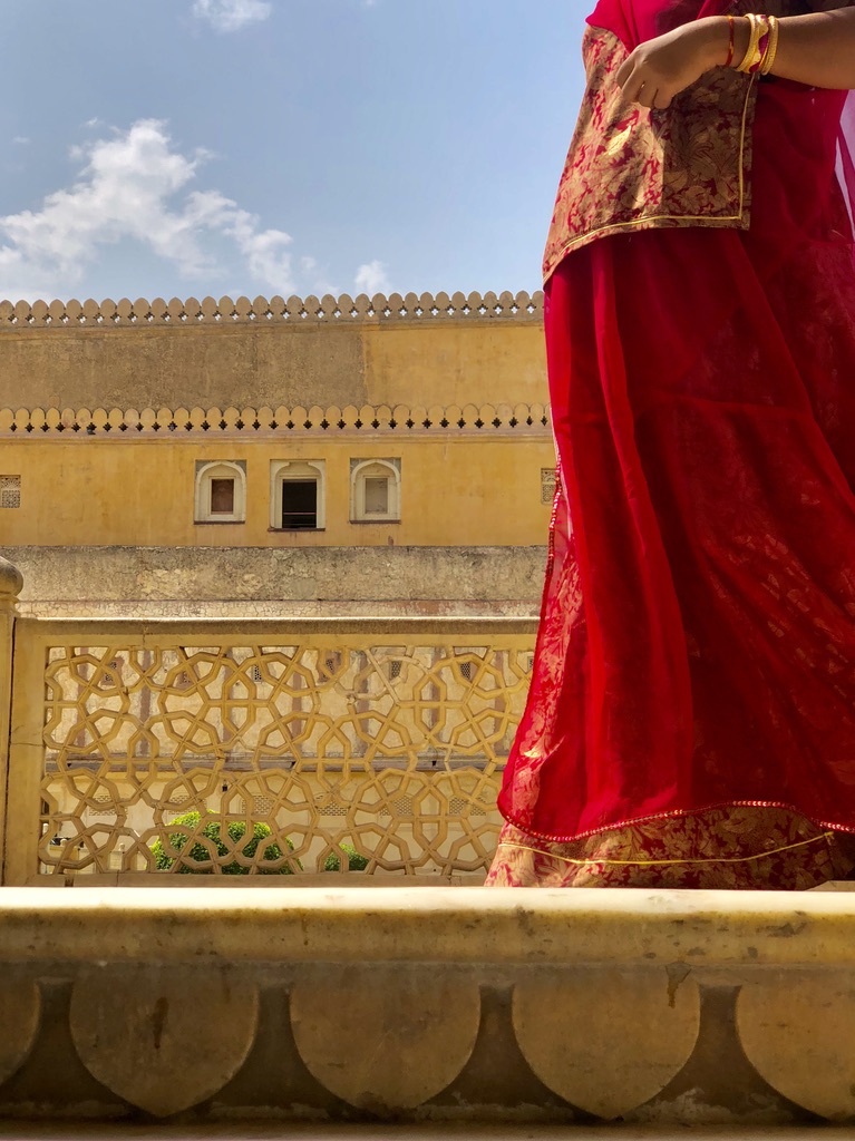 A person in a vibrant red and gold traditional outfit stands on a stone balcony with intricate latticework. The background features a yellow, historically styled building with decorative elements under a clear blue sky. The person's face is not visible.