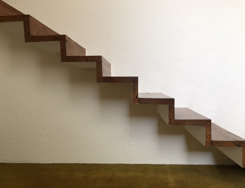 The image shows a modern, minimalist wooden staircase with a zigzag design against a plain white wall. The steps appear to be cantilevered, creating a floating effect, and there is no visible railing. The floor beneath is covered with a mustard-colored carpet.