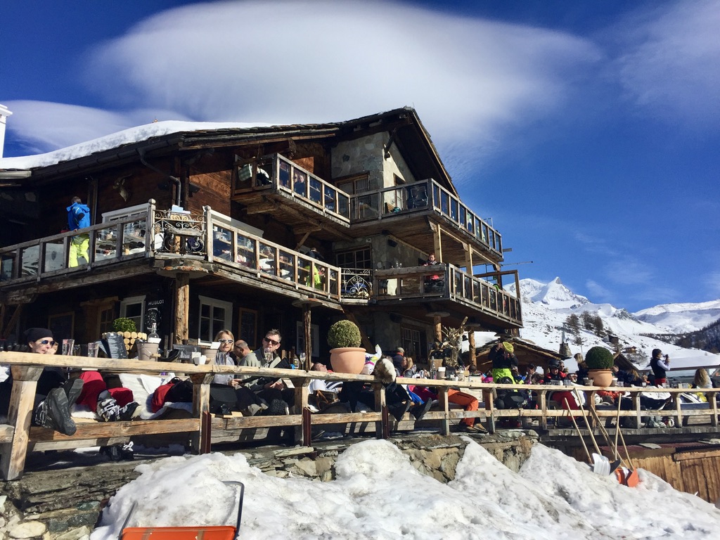 A rustic mountain lodge on a snowy slope with people sitting on the outdoor terrace, enjoying the sunny weather. The sky is clear blue with a unique cloud formation above. Snow-capped mountains are visible in the background.