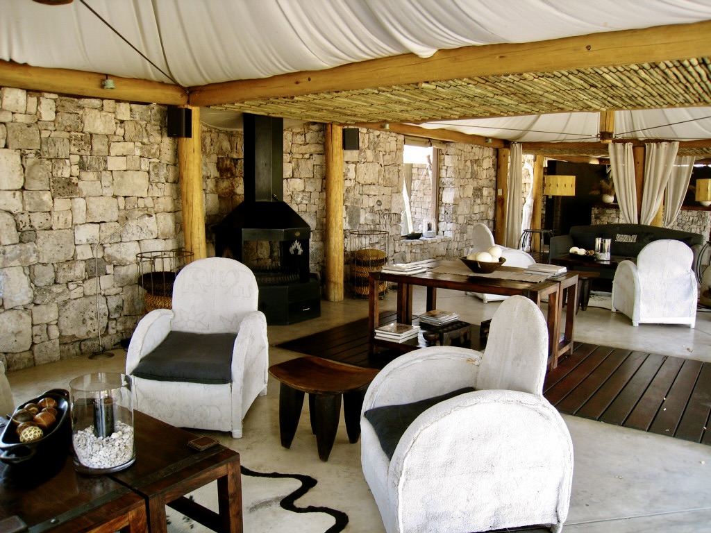 Elegant rustic lounge area with stone walls and a dark wooden floor, featuring white upholstered chairs, a wooden coffee table, and a central black fireplace under a white canvas ceiling.