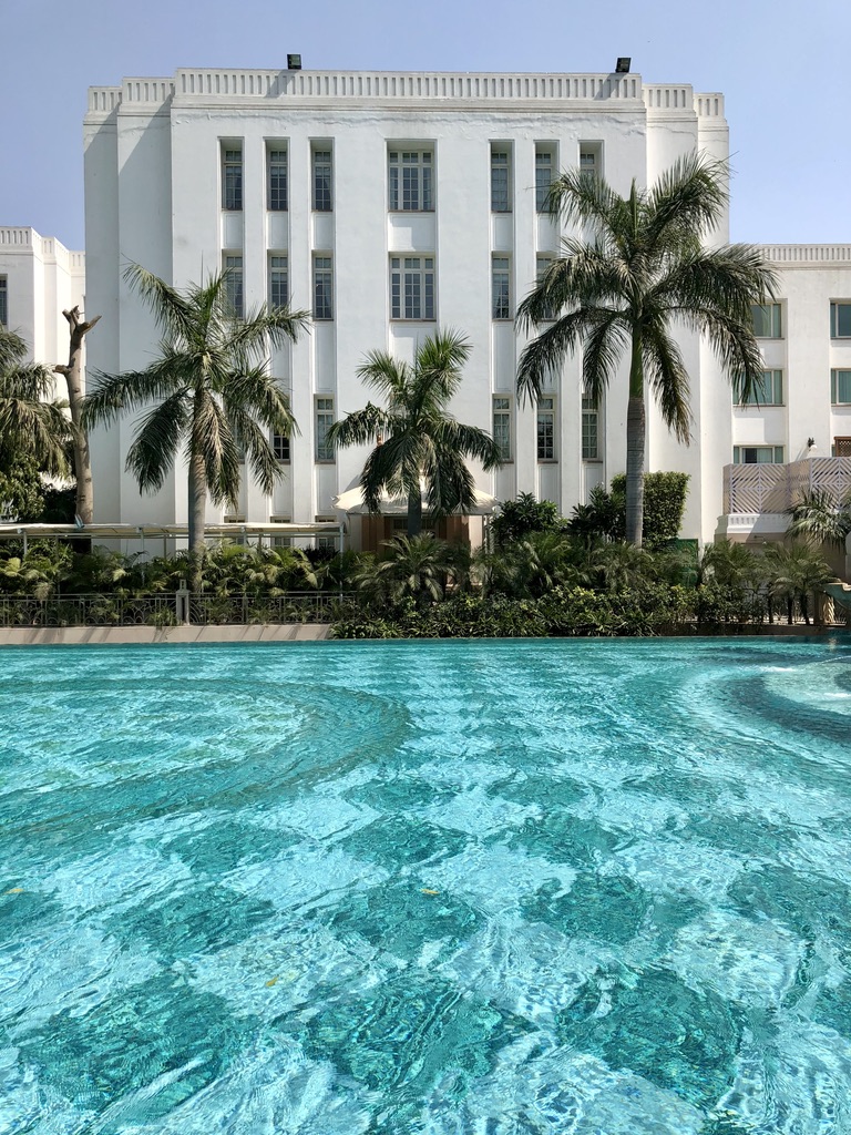 A large building with white walls stands behind a crystal-clear swimming pool. Palm trees and lush greenery line the edge of the pool, enhancing the tropical and luxurious feel of the setting. The sky is clear and blue, suggesting a sunny day.