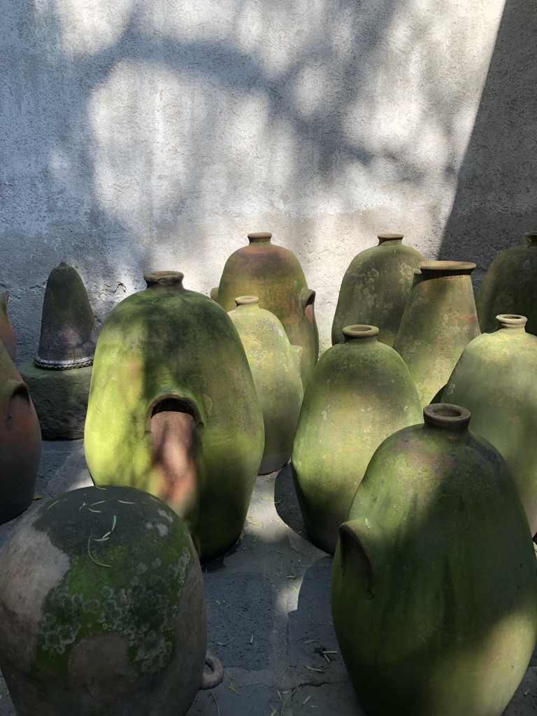 A collection of moss-covered, green ceramic pots of various sizes are set against a textured gray wall. Shadows from tree branches play on the wall and pots, creating an atmospheric, rustic scene. One pot has a hole near its base.