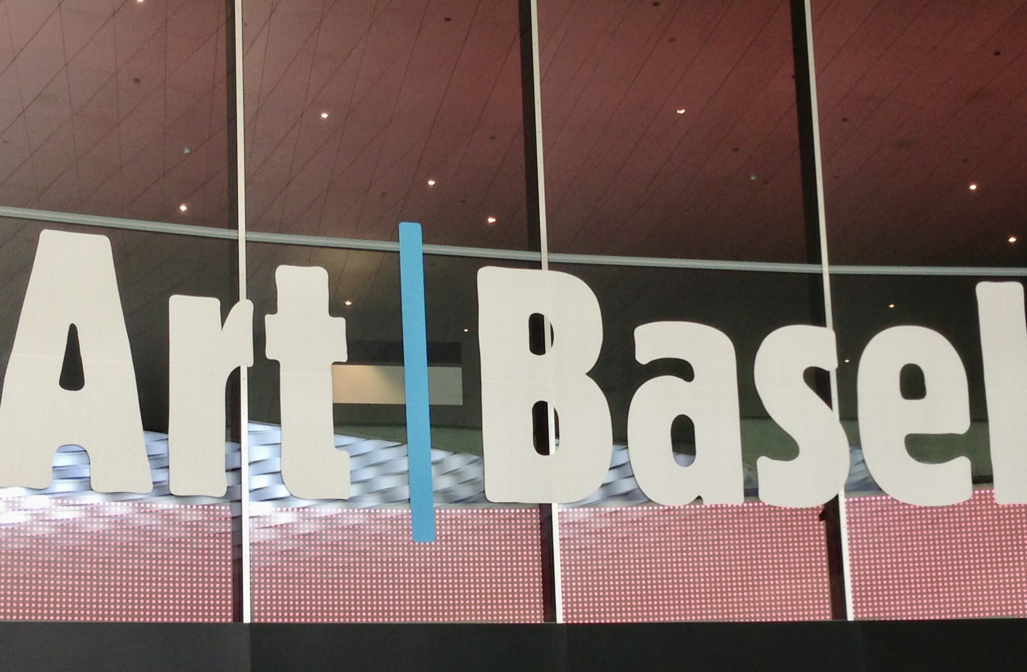 A sign reads "Art Basel" with large, bold white letters separated by a vertical blue line. The background features a reflective glass surface, showing a ceiling with a geometric pattern and a red dotted design near the bottom.