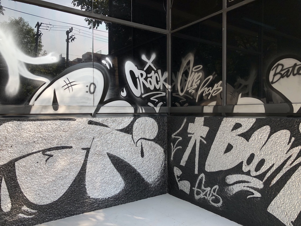 A building corner adorned with various graffiti tags and designs. The artwork includes large, bold letters in white and black spray paint, featuring phrases like "Crack," and "Boom." The textured wall and glass windows reflect the urban street art style.