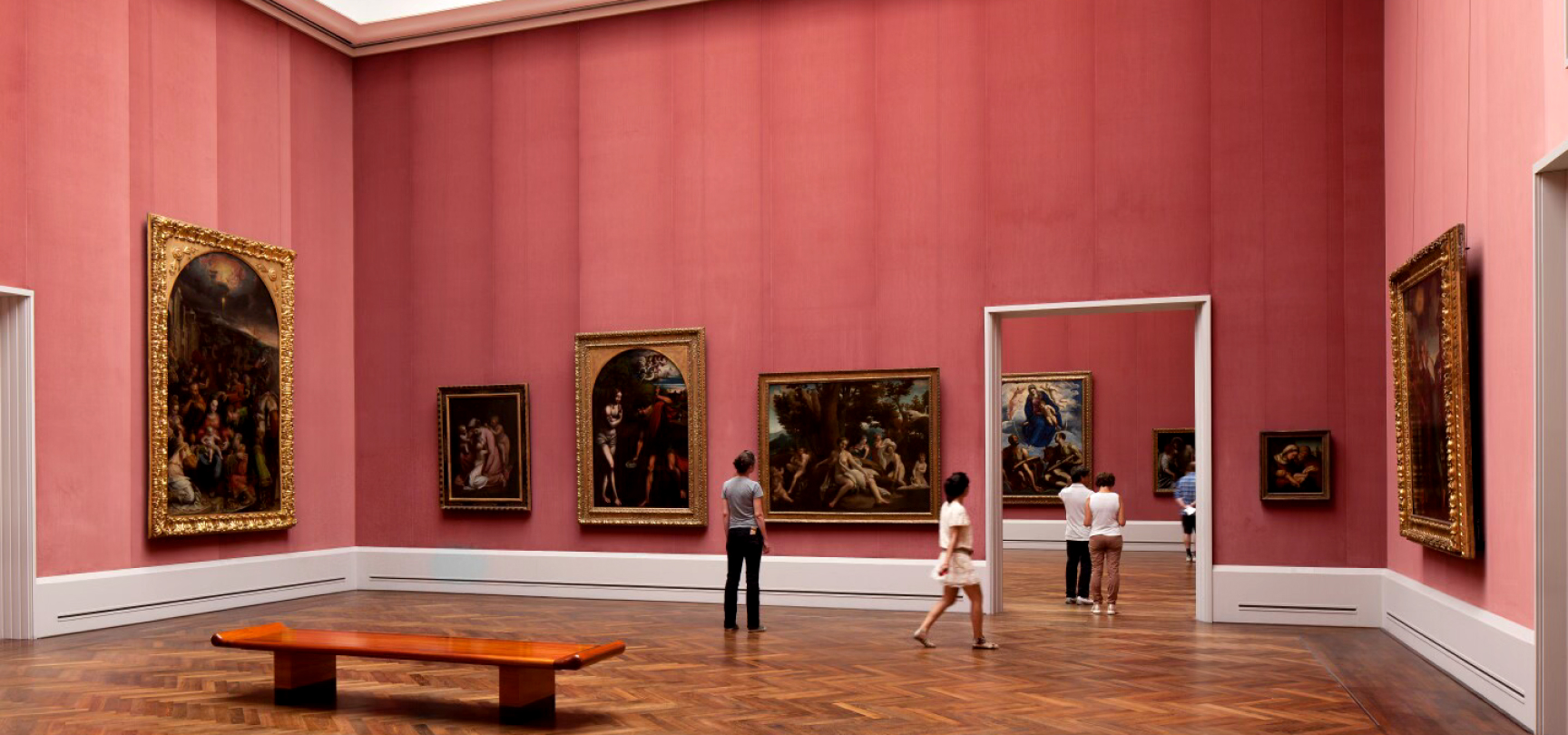 A spacious art gallery room with pink walls displaying various classical paintings in ornate gold frames. Several visitors are observing the artwork. The floor is parqueted wood, and there is a wooden bench in the foreground. Doorways lead to additional rooms.