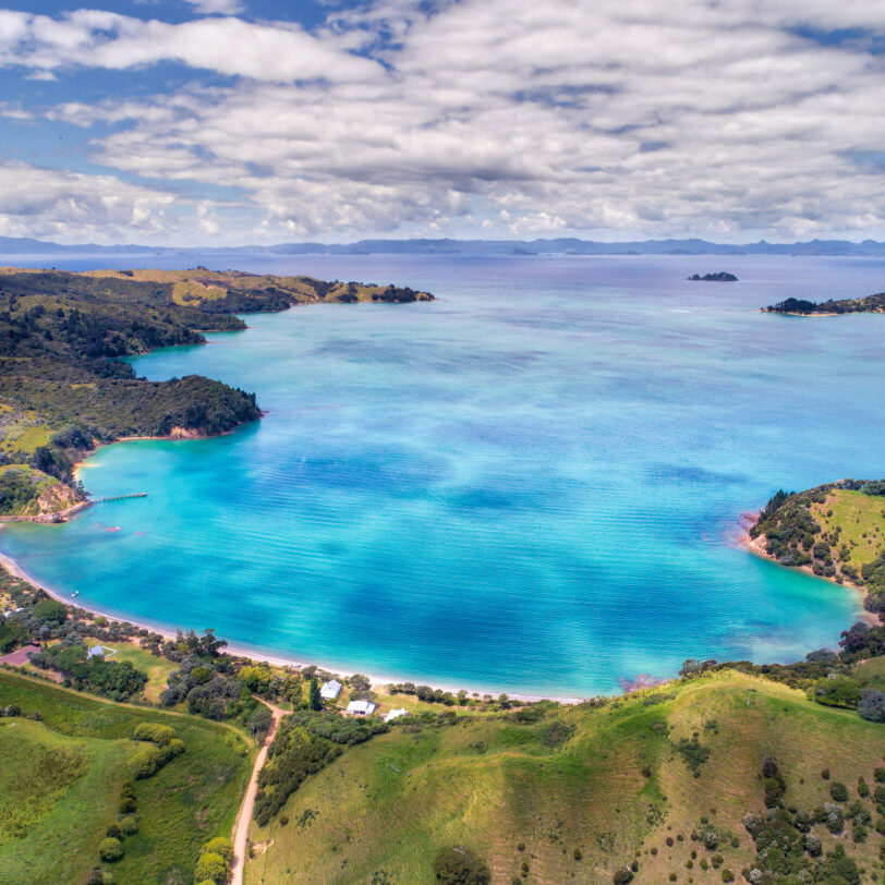 Aerial view of serene Wiheke coastal landscape featuring a turquoise bay surrounded by lush green hills and small islands in the distance. The sky is partly cloudy, adding depth to the vibrant scene. A few buildings and roads are visible near the shoreline.