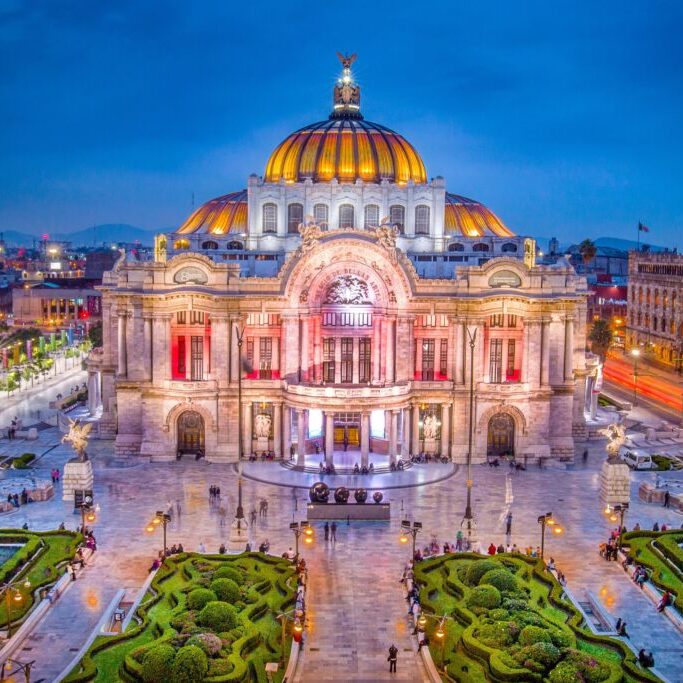 Aerial view of the Palacio de Bellas Artes (Palace of Fine Arts) in Mexico City, illuminated at dusk. The grand building features a distinctive yellow and orange dome, and the surrounding area includes neatly manicured gardens and bustling streets.