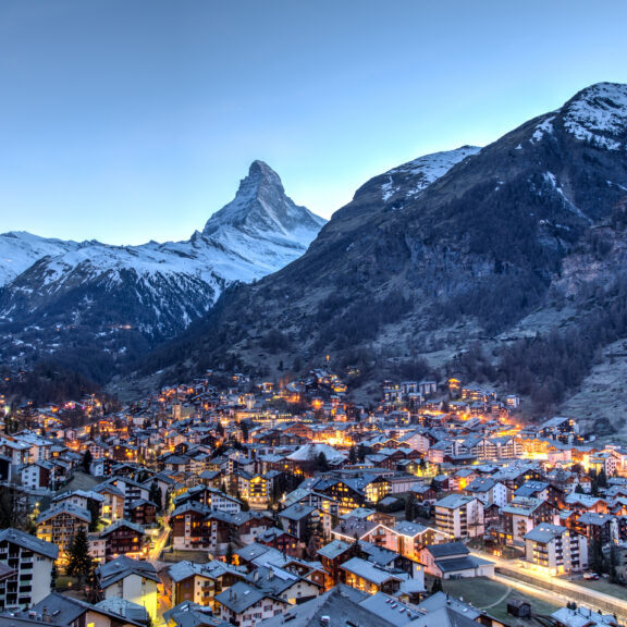 A picturesque mountain village at dusk, illuminated warmly against the backdrop of snow-capped peaks under a clear sky. The iconic Matterhorn mountain stands majestically in the background, towering over the clustered buildings below.