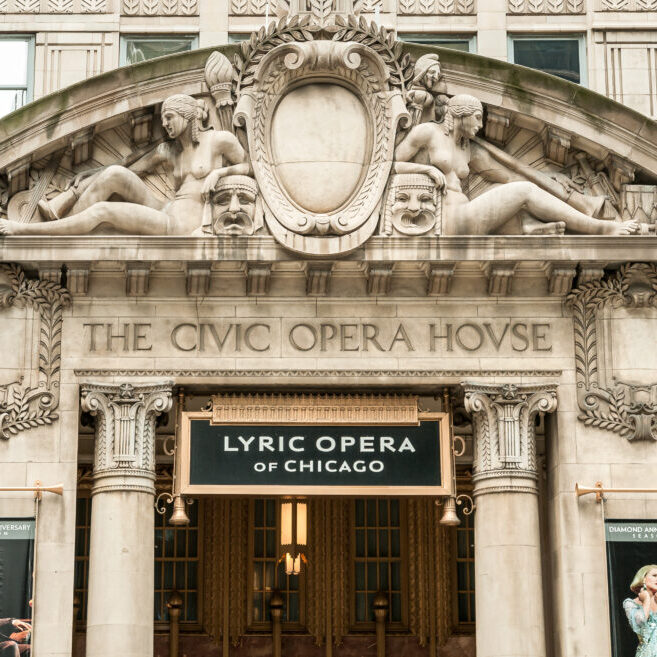 The ornate entrance of The Civic Opera House in Chicago, showcasing intricate sculptures and architectural details. Above the doors, a sign reads "Lyric Opera of Chicago," flanked by posters of opera performances.