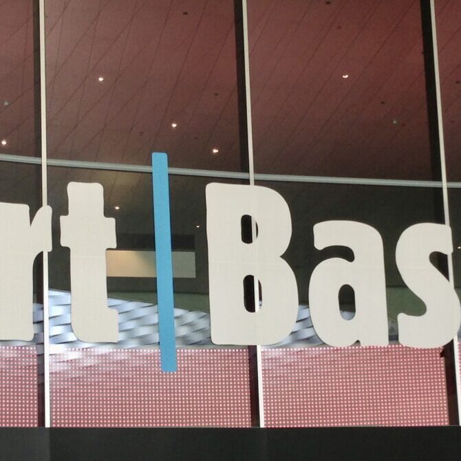 A sign reads "Art Basel" with large, bold white letters separated by a vertical blue line. The background features a reflective glass surface, showing a ceiling with a geometric pattern and a red dotted design near the bottom.