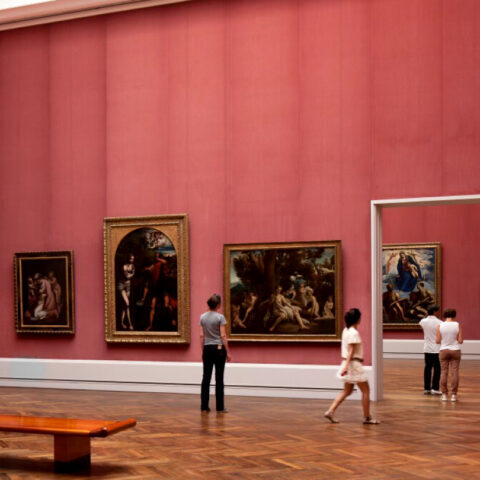 A spacious art gallery room with pink walls displaying various classical paintings in ornate gold frames. Several visitors are observing the artwork. The floor is parqueted wood, and there is a wooden bench in the foreground. Doorways lead to additional rooms.